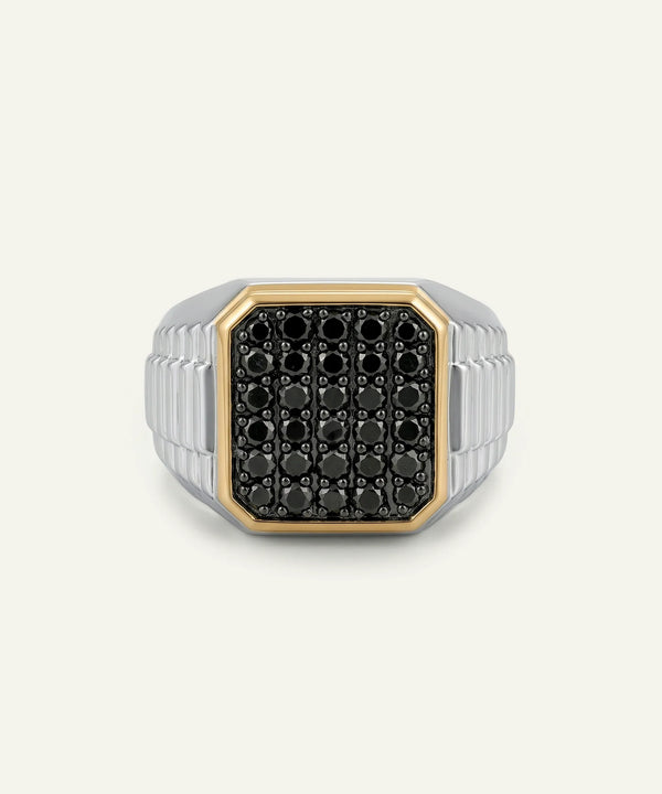 A men's signet ring featuring a black pavé face with a gold bezel, set on a silver-toned ribbed band, shown against a light off-white background.