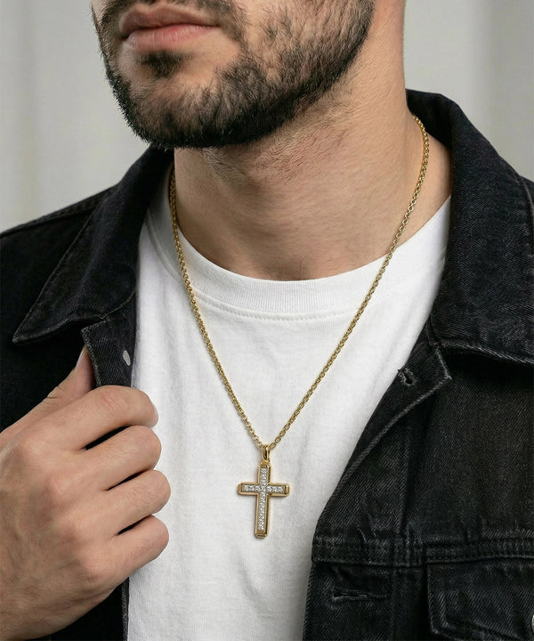 A close-up of a man wearing a gold cross pendant necklace with clear stones, styled over a white t-shirt and a dark denim jacket.
