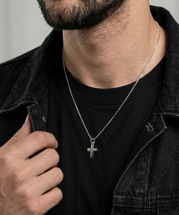 A close-up of a man wearing a silver cross pendant necklace over a black t-shirt and dark denim jacket.