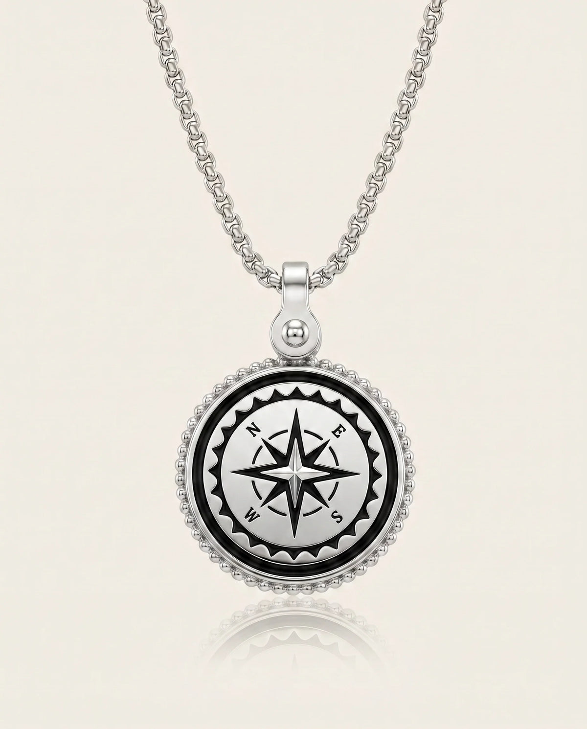 A silver compass pendant necklace on a matching chain, featuring a black compass rose design in the center with white lettering, surrounded by a beaded silver border, set against a light cream background.