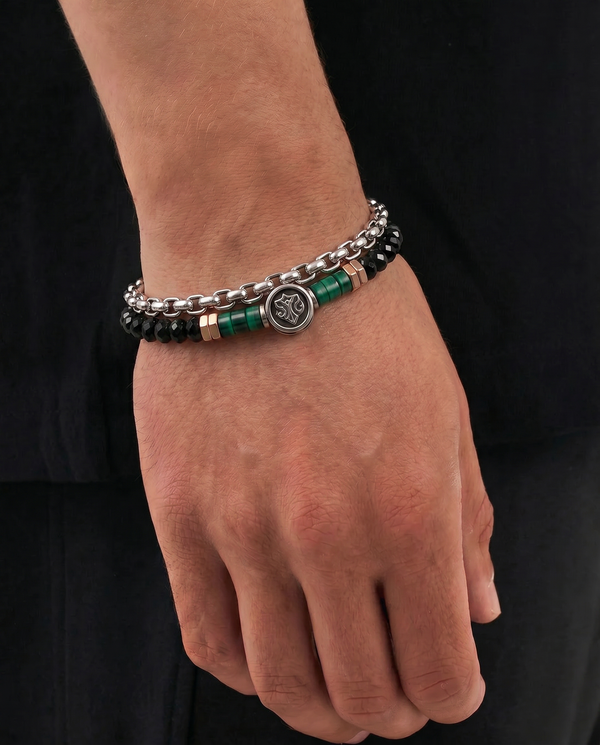 A close-up of a person's wrist wearing a double-strand bracelet. The bracelet features silver-tone chain links, black faceted beads, and polished green cylindrical beads separated by small gold-tone spacer rings, with a central round silver-tone charm.