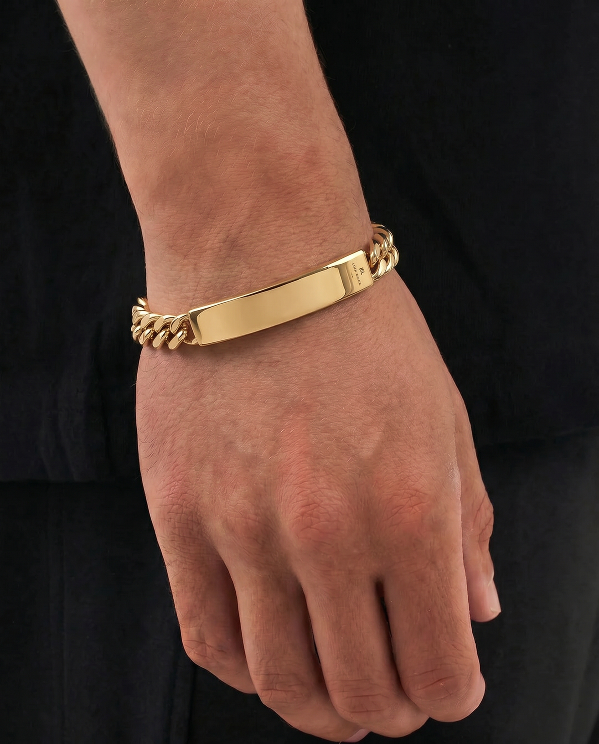 A gold-plated ID bracelet with a smooth central bar and curb-link ends, shown worn on a person's wrist against a black shirt background.