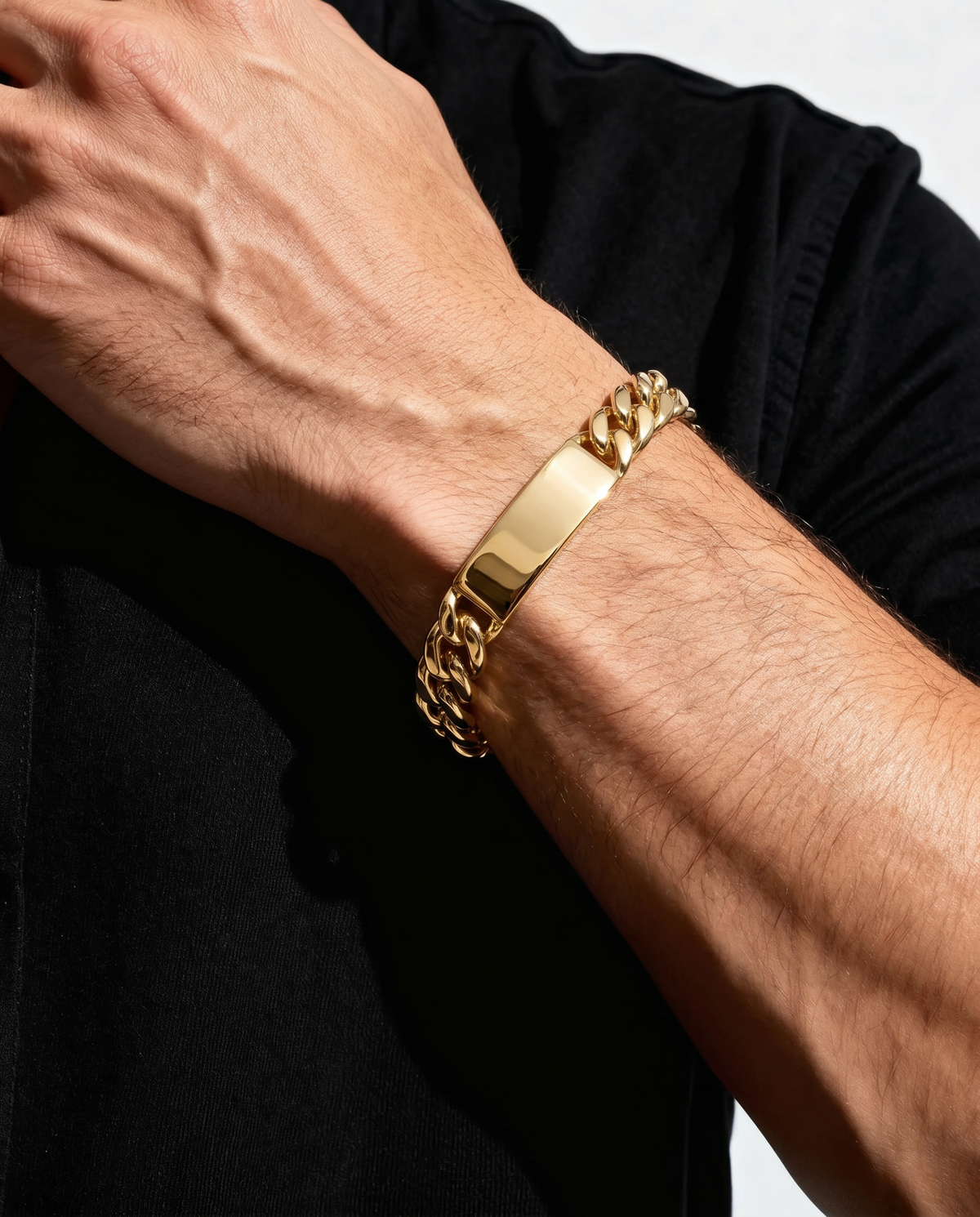 A close-up of a person's forearm and hand wearing a gold-tone Cuban link chain bracelet with a polished rectangular ID plate, paired with a black shirt.