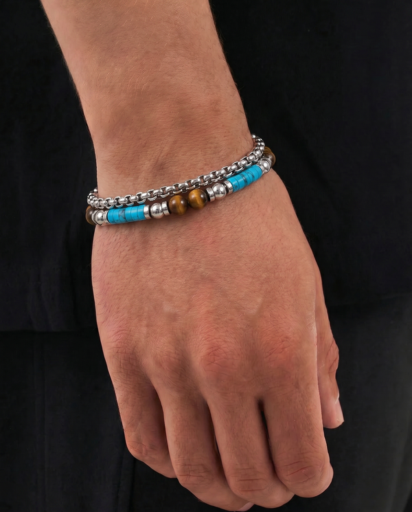 A close-up of a person's wrist and hand wearing two beaded bracelets. One bracelet is made of silver-toned metal beads, and the other features bright turquoise blue beads with silver spacer beads and a single brown tiger's eye bead.