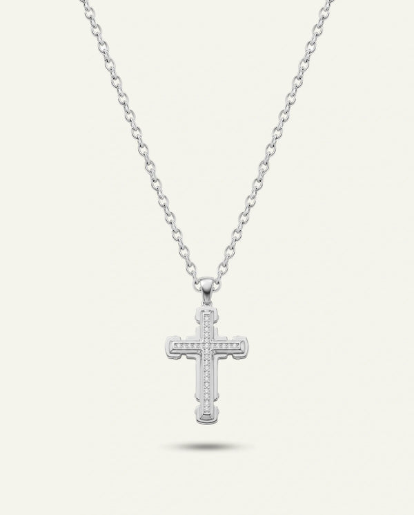 A silver cross pendant necklace with a delicate chain, shown against a light cream background with a soft shadow underneath.