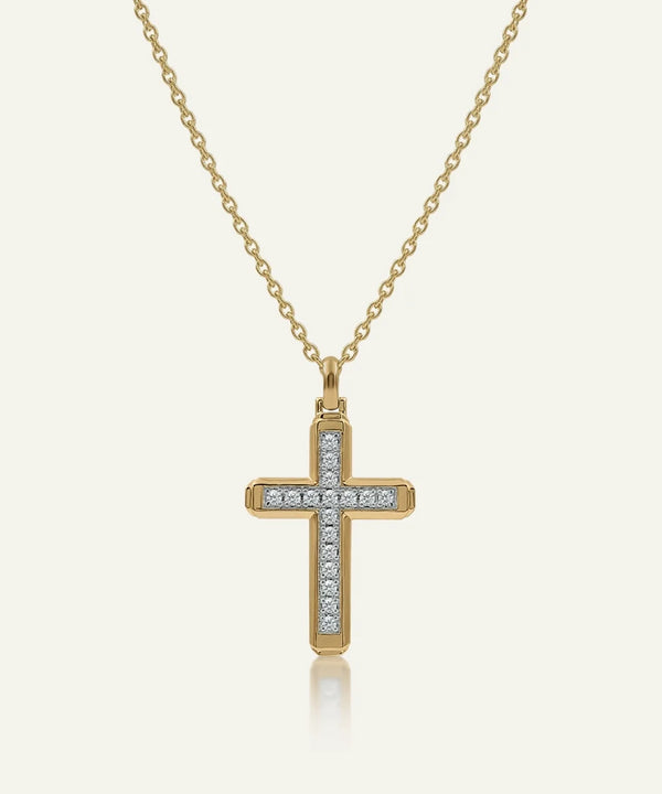 A gold cross pendant necklace adorned with small clear stones along the horizontal and vertical bars, suspended from a delicate gold chain against a light cream background.