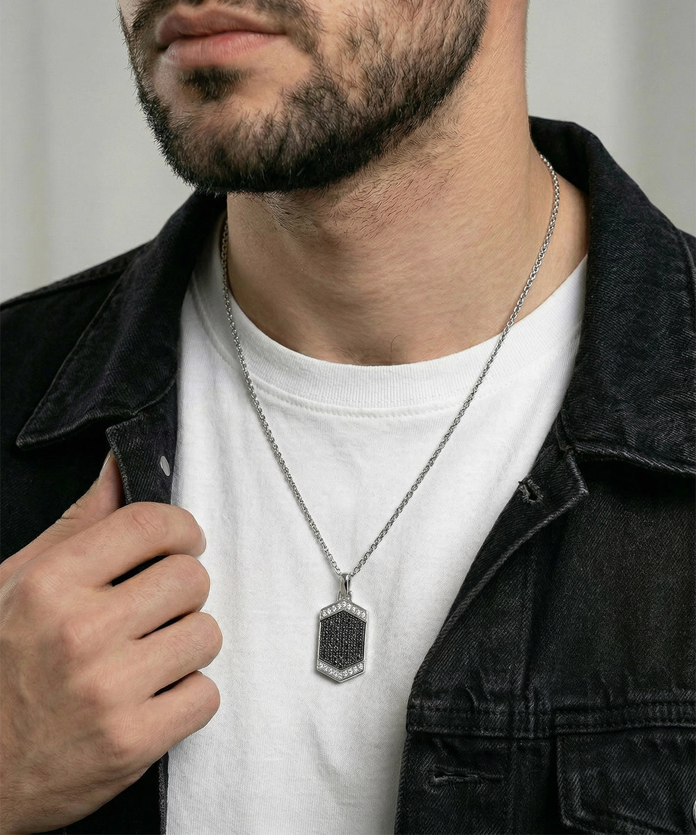 A close-up of a man wearing a silver-toned hexagonal pendant necklace with a black textured center, styled over a white t-shirt and a dark denim jacket.