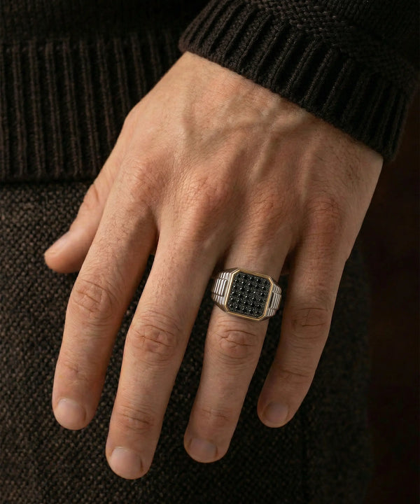 A person's hand wearing a men's two-tone silver and gold signet ring paved with black stones against a dark background.