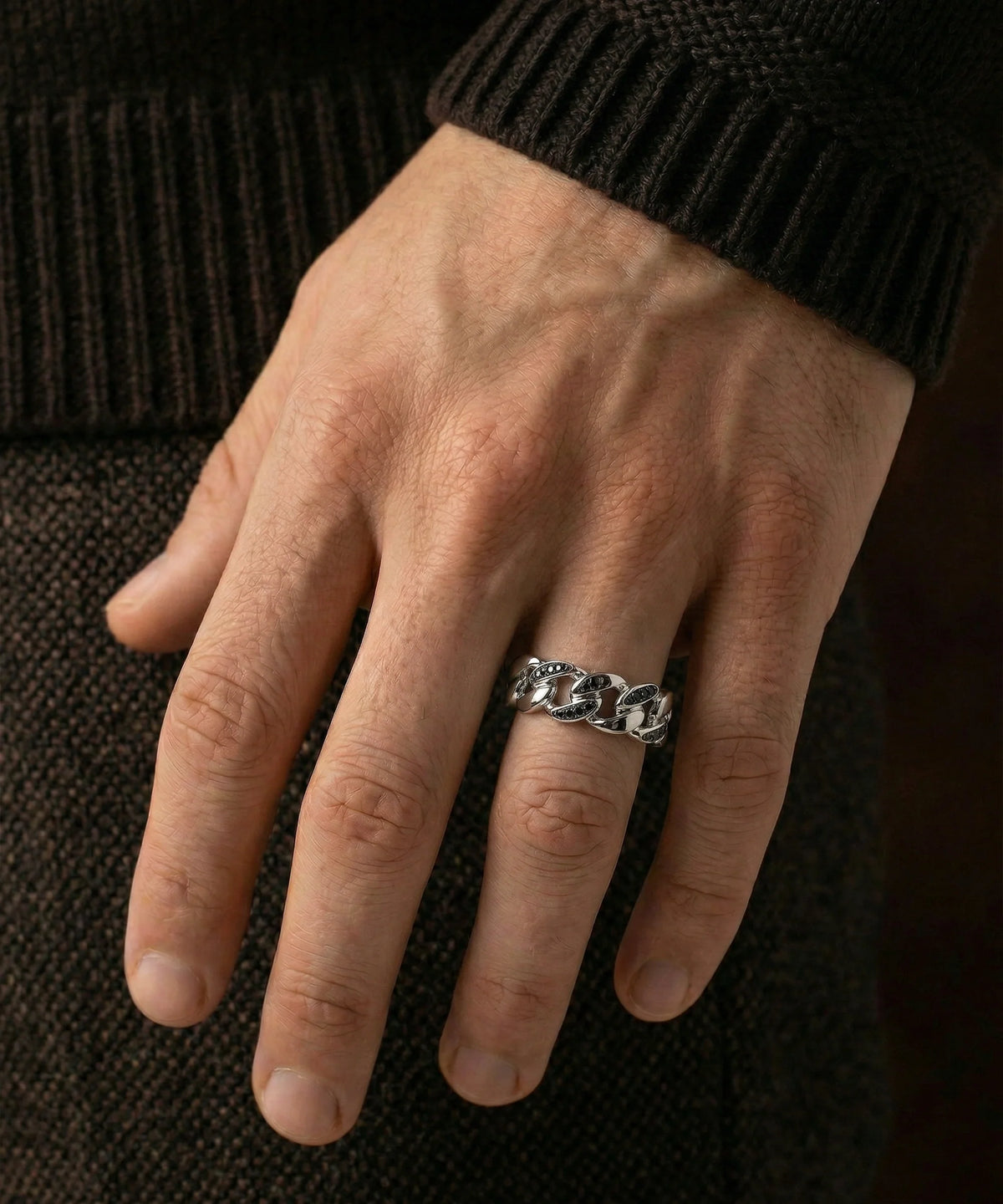 A person's hand wearing a men's silver Cuban link band ring set with clear and black stones against a dark background.