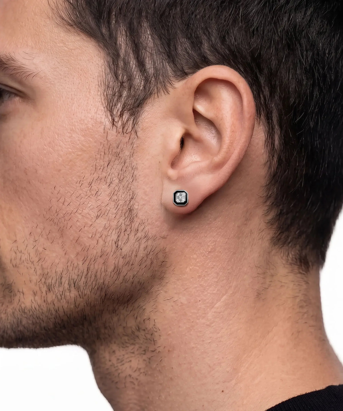 Close-up lifestyle shot of a man's side profile wearing a square silver stud earring with a dark center stone set in a black halo border.