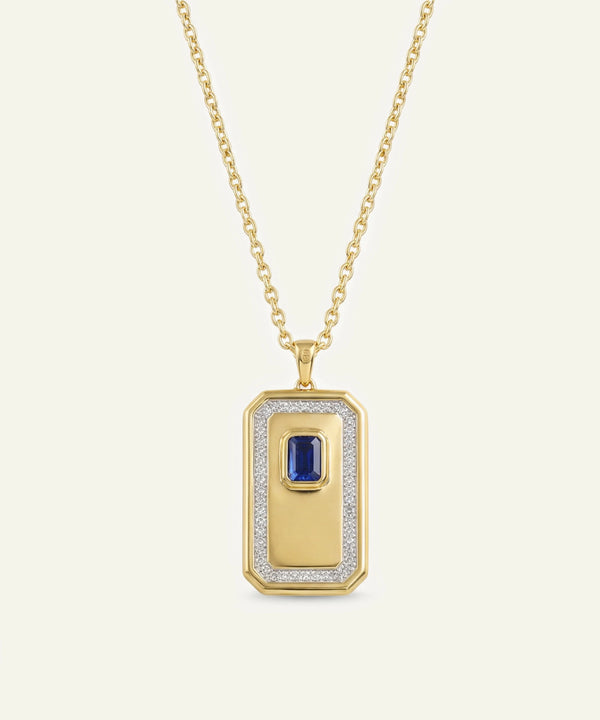 A gold pendant necklace featuring a rectangular dog tag-style pendant with a central blue sapphire and a halo of small clear stones, shown on a light cream background.