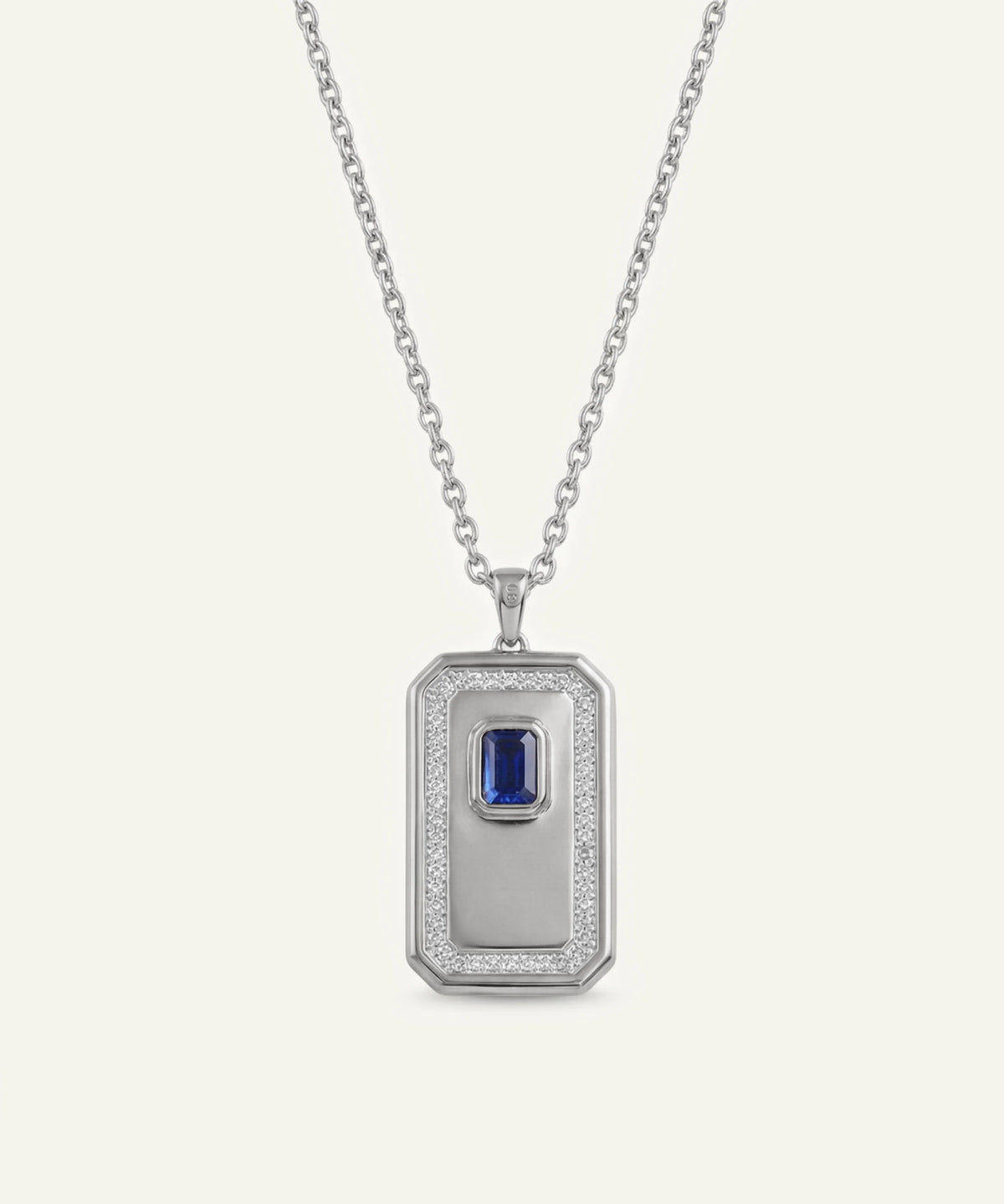 A silver-toned rectangular pendant necklace featuring a central blue sapphire gemstone surrounded by a halo of small clear stones, suspended from a delicate silver chain against a light cream background.
