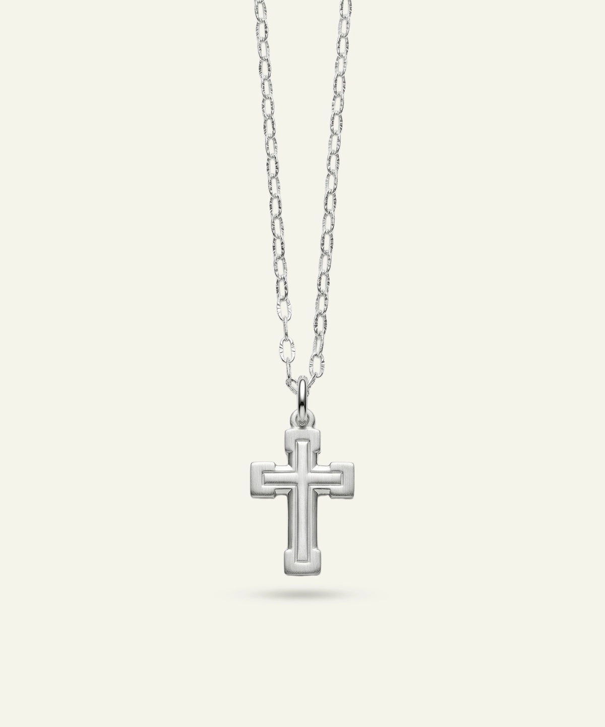 A silver cross pendant necklace with a textured, raised geometric border design, suspended from a delicate silver link chain against a light cream background.