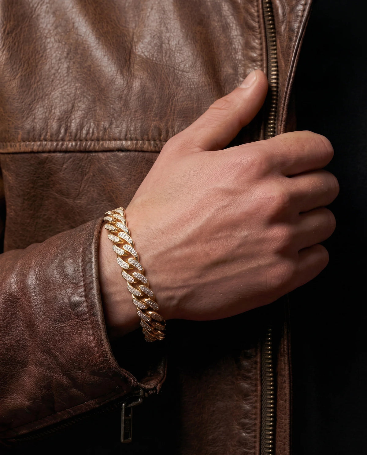 Close-up lifestyle shot of a man's wrist wearing a gold-toned pavé diamond-encrusted Miami Cuban chain bracelet, styled against a brown leather jacket.