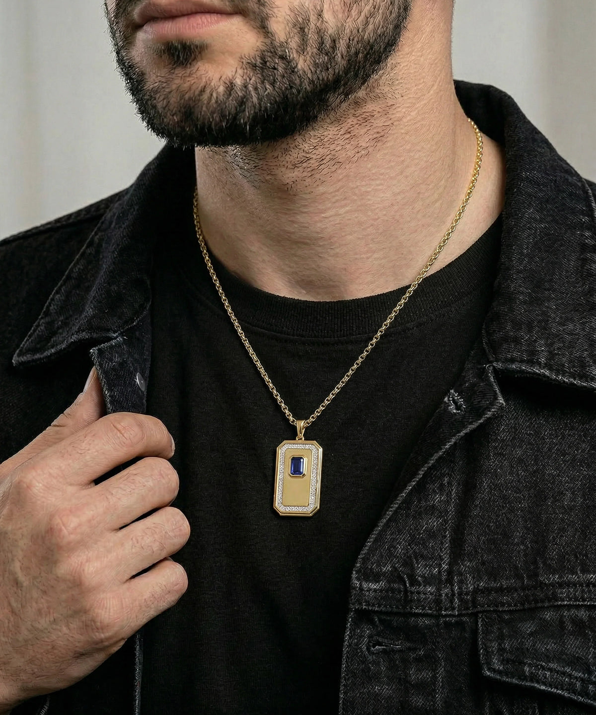 A close-up of a man wearing a gold rectangular pendant necklace with a blue and white inlay, styled over a black t-shirt and dark denim jacket.