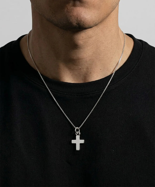 A silver cross pendant necklace worn by a person in a black crewneck t-shirt against a light gray background.
